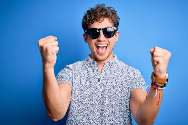 Genç sarışın, kıvırcık saçlı, tatilde çizgili polo ve güneş gözlüğü takan yakışıklı adam kolları açık ve kolları açık bir şekilde başarıyı kutluyor ve şaşırıyor. Kazanan konsept.