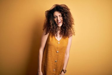 Beautiful brunette woman on vacation with curly hair and piercing wearing hat and dress winking looking at the camera with sexy expression, cheerful and happy face.