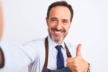 Önlük giyen orta yaşlı bir esnaf izole edilmiş beyaz arka planda selfie çeker. Gülümseyerek mutlu olur. İşaret yapar.