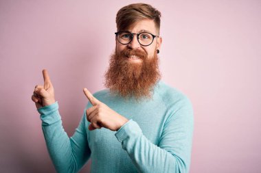 Pembe izole edilmiş arka planda gözlük takan, sakallı, yakışıklı kızıl saçlı, gülümseyen ve kameraya bakan, iki eli ve parmakları yanda olan yakışıklı bir adam..