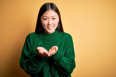 Young beautiful asian woman wearing green winter sweater over yellow isolated background Smiling with hands palms together receiving or giving gesture. Hold and protection
