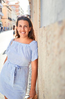 Genç ve güzel bir kadın mutlu bir şekilde gülümsüyor güneşli bir yaz gününde şehir sokaklarının duvarına yaslanıyor.