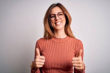Genç, güzel, esmer bir kadın beyaz arka plan işareti üzerine günlük süveter ve gözlük takıyor. Elleriyle olumlu jest yapıyor. Gülümsüyor ve mutlu. Neşeli ifade ve kazanan jest.