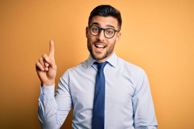 Young handsome businessman wearing tie and glasses standing over yellow background pointing finger up with successful idea. Exited and happy. Number one.