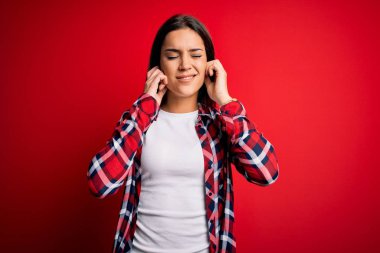 Young beautiful brunette woman wearing casual shirt standing over isolated red background covering ears with fingers with annoyed expression for the noise of loud music. Deaf concept.