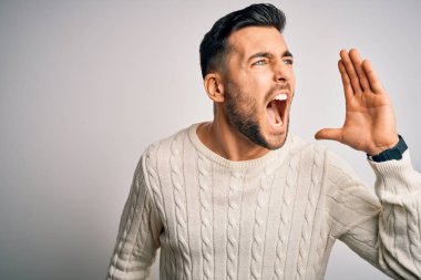 Young handsome man wearing casual sweater standing over isolated white background shouting and screaming loud to side with hand on mouth. Communication concept.