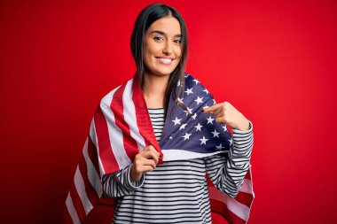 Young beautiful brunette patriotic woman wearing united states flag for independence day with surprise face pointing finger to himself
