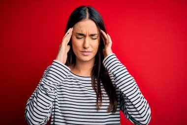 Young beautiful brunette woman wearing casual striped t-shirt over red background with hand on head for pain in head because stress. Suffering migraine.