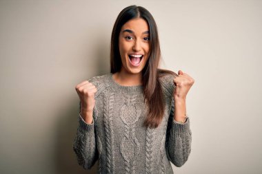 Young beautiful brunette woman wearing casual sweater over isolated white background celebrating surprised and amazed for success with arms raised and open eyes. Winner concept.