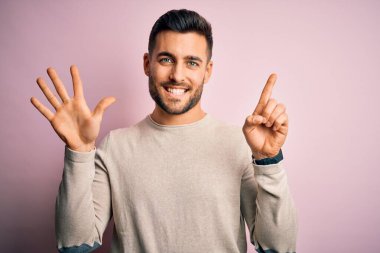 Young handsome man wearing casual sweater standing over isolated pink background showing and pointing up with fingers number six while smiling confident and happy.