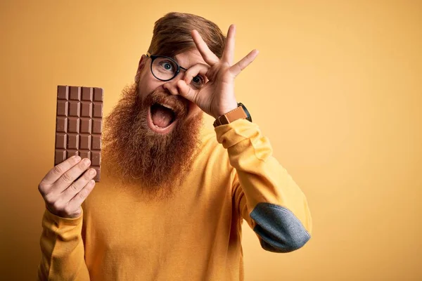 Irish redhead man with beard eating a sweet chocolate bar over yellow isolated background with happy face smiling doing ok sign with hand on eye looking through fingers