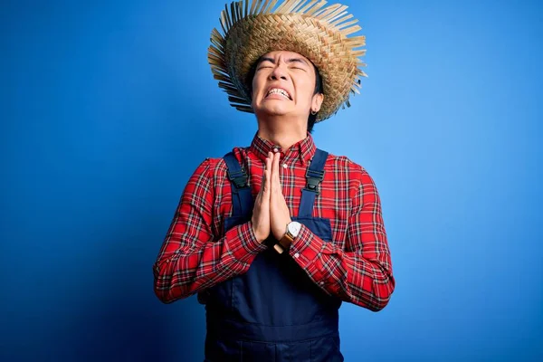 Young handsome chinese farmer man wearing apron and straw hat over blue background begging and praying with hands together with hope expression on face very emotional and worried. Begging.