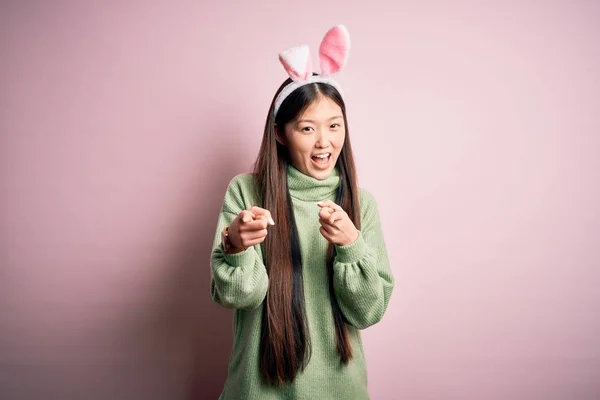 年轻的亚洲女人 戴着可爱的复活节兔子耳朵 粉红的背景 手指指向相机 脸上带着快乐和滑稽的表情 好的能量和活力 — 图库照片