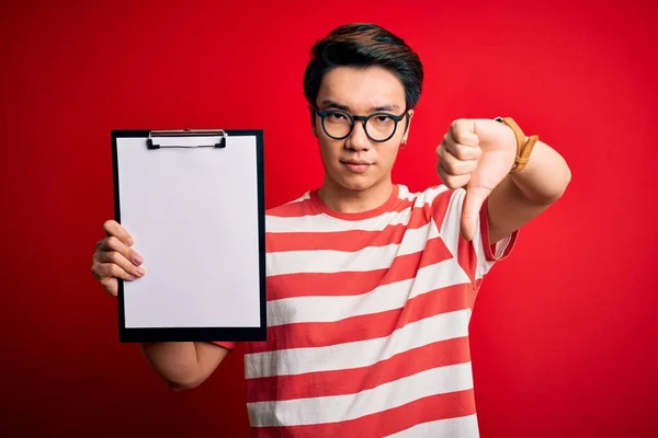 Young handsome chinese inspector man wearing glasses holding clipboard checklist with angry face, negative sign showing dislike with thumbs down, rejection concept