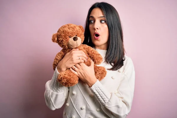 Young brunette woman with blue eyes hugging teddy bear stuffed animal over pink background scared in shock with a surprise face, afraid and excited with fear expression