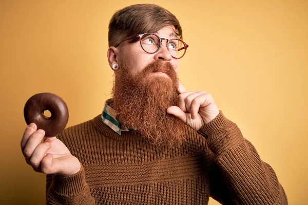 Redhead Irish man with beard eating chocolate donut over yellow background serious face thinking about question, very confused idea