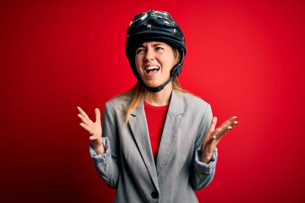 若い美しいブロンドのオートバイの女性は 赤い背景の狂気と怒りの叫びにオートバイのヘルメットをかぶって積極的な表情と腕を上げて叫びました 欲求不満の概念 — ストック写真