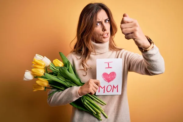 Beautiful brunette woman holding love mom message and tulips celebrating mothers day annoyed and frustrated shouting with anger, crazy and yelling with raised hand, anger concept
