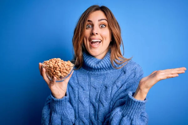 Young Beautiful Brunette Woman Holding Bowl Peanuts Blue Background Very — Stock Photo, Image