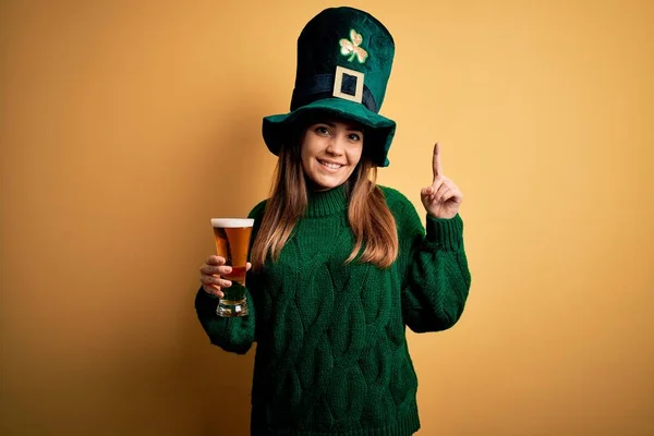 顔に大きな笑顔で聖人の公園の日にビールの緑の帽子を飲むガラスを身に着けている若い美しい女性 カメラを見て側に手指で指して — ストック写真