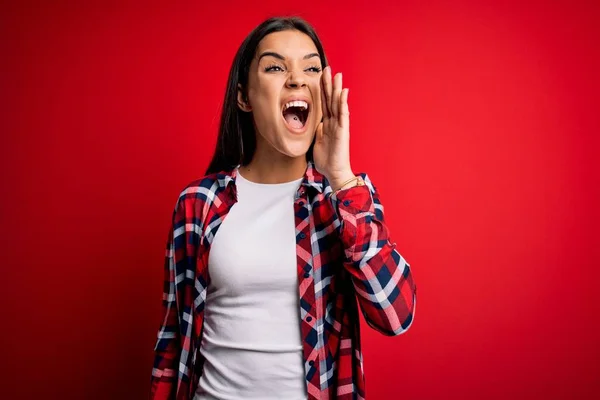 Young beautiful brunette woman wearing casual shirt standing over isolated red background shouting and screaming loud to side with hand on mouth. Communication concept.