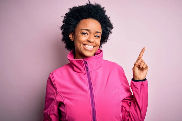 Young African American afro sportswoman with curly hair wearing sportswear doin sport with a big smile on face, pointing with hand and finger to the side looking at the camera.