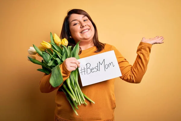 Beautiful plus size woman celebrating mothers day holding best mom message and tulips very happy and excited, winner expression celebrating victory screaming with big smile and raised hands