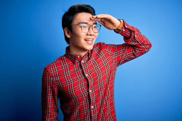 Young handsome chinese man wearing casual shirt and glasses over blue background very happy and smiling looking far away with hand over head. Searching concept.