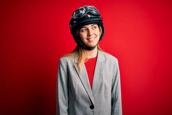 若い美しいブロンドのオートバイの女性の顔に笑顔で横に離れて見て赤い背景にオートバイのヘルメットを着て 自然な表情 自信を持って — ストック写真