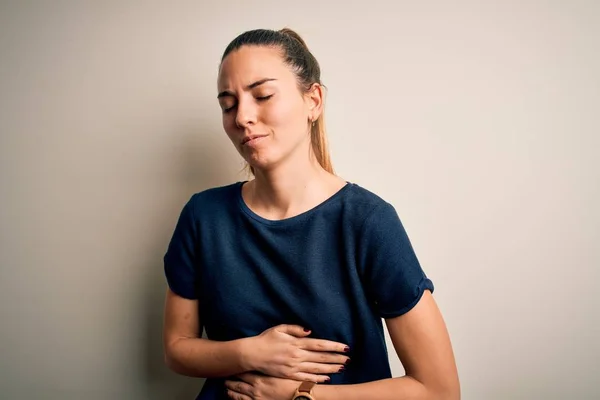 胃の上に手で白い背景にカジュアルなTシャツを着て青い目を持つ若い美しいブロンドの女性ので 消化不良 痛みを伴う病気の気分が悪い アチェの概念 — ストック写真