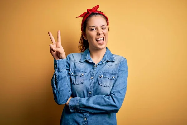 Young beautiful brunette woman wearing casual denim shirt and hair handkerchief smiling with happy face winking at the camera doing victory sign. Number two.