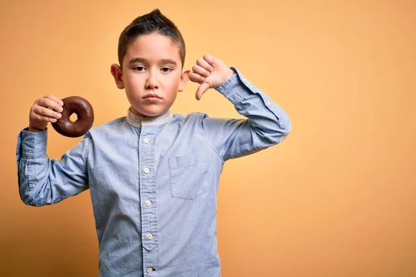 若い男の子食べる不健康なチョコレートドーナツ上の隔離された黄色の背景で怒っている顔 否定的な兆候ショー嫌いで親指ダウン 拒否の概念 — ストック写真