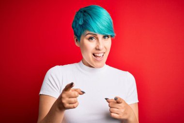 Young beautiful woman with blue fashion hair wearing casual t-shirt over red background pointing fingers to camera with happy and funny face. Good energy and vibes.