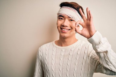 Young handsome chinese man injured for accident wearing bandage and strips on head smiling positive doing ok sign with hand and fingers. Successful expression.