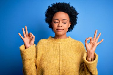 Kıvırcık saçlı, sarı, rahat kazak giyen, gözleri kapalı gülümseyen, meditasyon hareketi yapan güzel Afro-Amerikan kadın. Yoga konsepti.