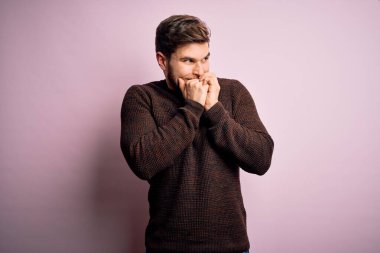Young blond man with beard and blue eyes wearing casual sweater over pink background laughing nervous and excited with hands on chin looking to the side