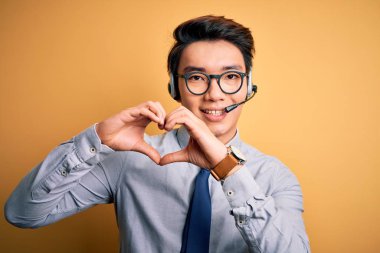 Young handsome chinese call center agent man wering glasses using headset smiling in love showing heart symbol and shape with hands. Romantic concept.