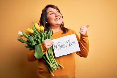 Beautiful plus size woman celebrating mothers day holding best mom message and tulips pointing and showing with thumb up to the side with happy face smiling