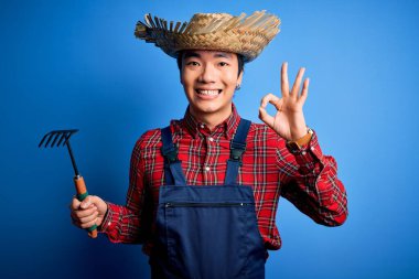 Young handsome chinese farmer man wearing apron and straw hat holding rake tool doing ok sign with fingers, excellent symbol