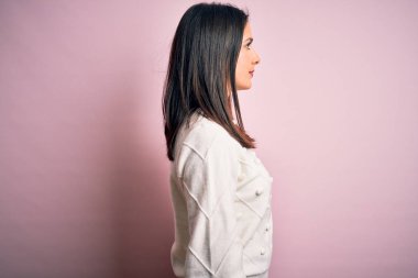 Mavi gözlü genç esmer kadın izole edilmiş pembe arka planda günlük süveter giyiyor, yan yana bakıyor, rahat poz veriyor, kendine güvenen bir gülüşü var..