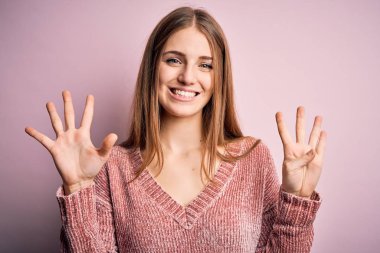 Genç, güzel, kızıl saçlı bir kadın pembe arka plan üzerine günlük süveter giyip dokuz numaralı parmaklarıyla işaret ederken kendinden emin ve mutlu bir şekilde gülümsüyor..