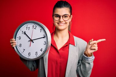 Young beautiful brunette businesswoman doing countdown holding big clock very happy pointing with hand and finger to the side