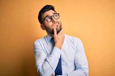 Sarı arka plan üzerinde duran kravat ve gözlük takan genç yakışıklı işadamı bir soru hakkında endişeli ve gergin bir şekilde düşünüyor.