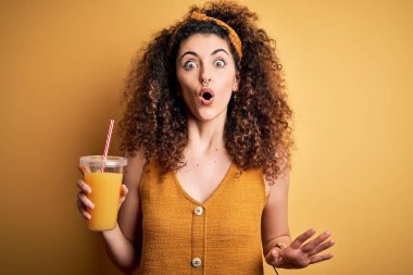 Young beautiful woman with curly hair and piercing drinking healthy orange juice scared in shock with a surprise face, afraid and excited with fear expression