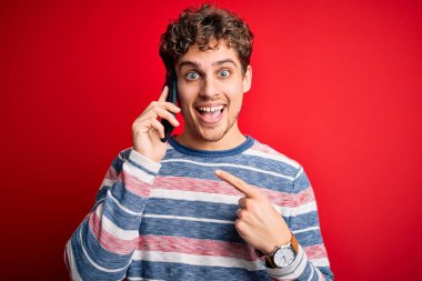 Sarışın, kıvırcık saçlı genç adam akıllı telefonla konuşuyor. El ve parmağıyla işaret ederek çok mutlu.