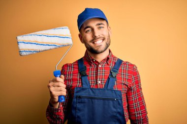 Young handsome painter man painting wall using roller over isolated yellow background with a happy face standing and smiling with a confident smile showing teeth