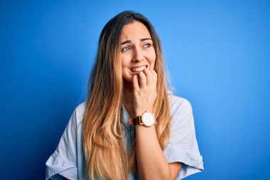 Young beautiful blonde woman with blue eyes wearing striped shirt over blue background looking stressed and nervous with hands on mouth biting nails. Anxiety problem.