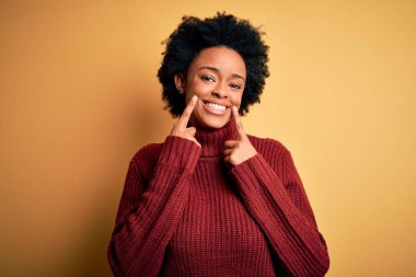 Kıvırcık saçlı, balıkçı yaka kazak giyen, ağzı açık gülümseyen, parmakları işaret eden ve neşeli gülümsemeye zorlayan güzel Afro-Amerikan kadın.