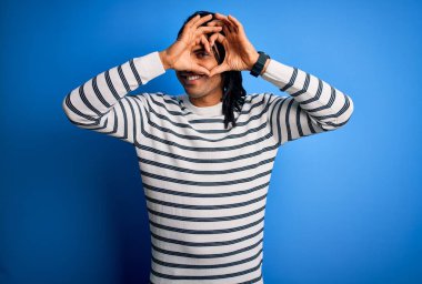 Young handsome african american afro man with dreadlocks wearing casual striped sweater Doing heart shape with hand and fingers smiling looking through sign