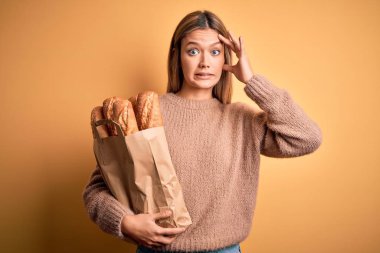 Young beautiful woman holding paper bag with bread over isolated yellow background stressed with hand on head, shocked with shame and surprise face, angry and frustrated. Fear and upset for mistake.